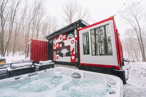 A snow-covered outdoor hot tub area with a red and white RV-style building, surrounded by snow and trees. - The Buckeye Box - The James (McArthur)