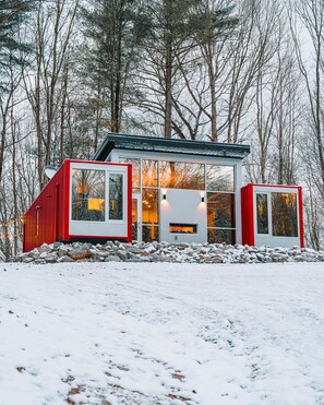 A modern cabin with large windows and a red exterior, surrounded by snow and trees. - The Buckeye Box - The James (McArthur)