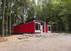 Une cabane rouge avec un toit noir, entourée d’arbres, et un chemin de gravier menant à celle-ci.