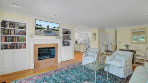 A spacious living room with a fireplace, a television mounted above it, and a bookshelf filled with books. There is a dining area with a table and chairs, and a kitchen in the background. - 43 Fairway Dunes by Avantstay Coastal Charm + Golf! (Isle of Palms)
