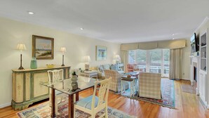 A spacious living room with a glass coffee table, a sofa, and a dining area with a table and chairs. There are large windows with curtains, a fireplace, and a television mounted on the wall. - 43 Fairway Dunes by Avantstay Coastal Charm + Golf! (Isle of Palms)