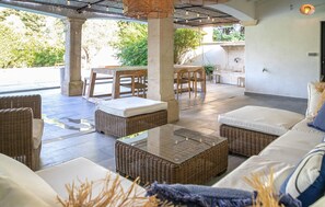 Un espacio de descanso al aire libre cubierto, con mobiliario de mimbre y una mesa de centro de vidrio.