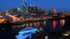 Un paisaje urbano nocturno con un río reflejando las luces de la ciudad, un edificio destacado con aguja, y un muelle con un cartel que dice "Nashville Riverfront".