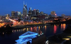 Un paisaje urbano nocturno con un río reflejando las luces de la ciudad, un edificio destacado con aguja, y un muelle con un cartel que dice "Nashville Riverfront".