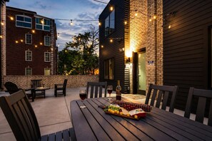 Une terrasse extérieure avec une table dressée pour un repas, entourée de chaises et illuminée par des guirlandes lumineuses suspendues au-dessus.