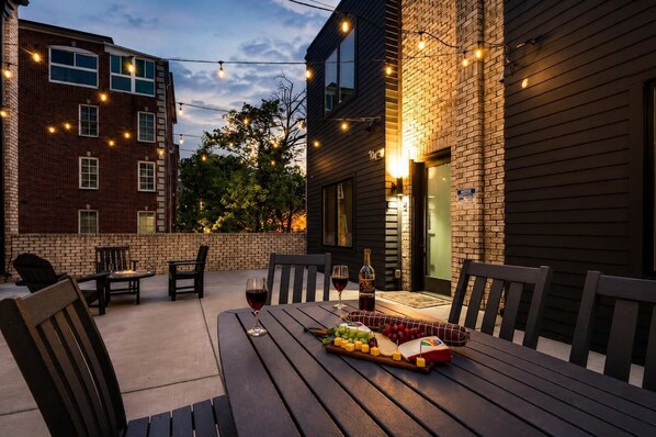 Een terras buiten met een eettafel, omringd door stoelen, en sfeerverlichting erboven.