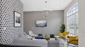 Una sala de estar moderna con un sofá gris, un sillón amarillo, otro sillón gris, un reposapiés azul, una alfombra gris, una araña y un ventanal grande con persianas.
