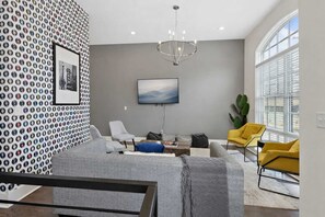 Una sala de estar moderna con un sofá gris, un sillón amarillo, otro sillón gris, un reposapiés azul, una alfombra gris, una araña y un ventanal grande con persianas.