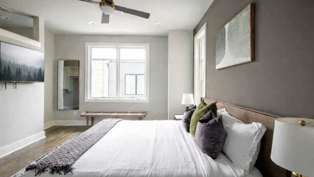 Un dormitorio con cama, ventilador de techo, ventana, banco y un cuadro en la pared.