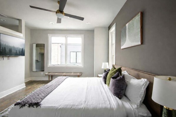 Un dormitorio con cama, ventilador de techo, ventana, banco y un cuadro en la pared.