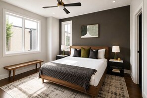 Une chambre à coucher avec un lit, des tables de chevet, un banc, un ventilateur de plafond et une fenêtre.