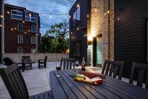 Une terrasse extérieure avec une table dressée pour un repas, entourée de chaises et illuminée par des guirlandes lumineuses suspendues au-dessus.