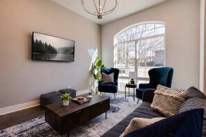 A living room with a large window, a dark sofa, a blue armchair, a coffee table, and a painting on the wall.