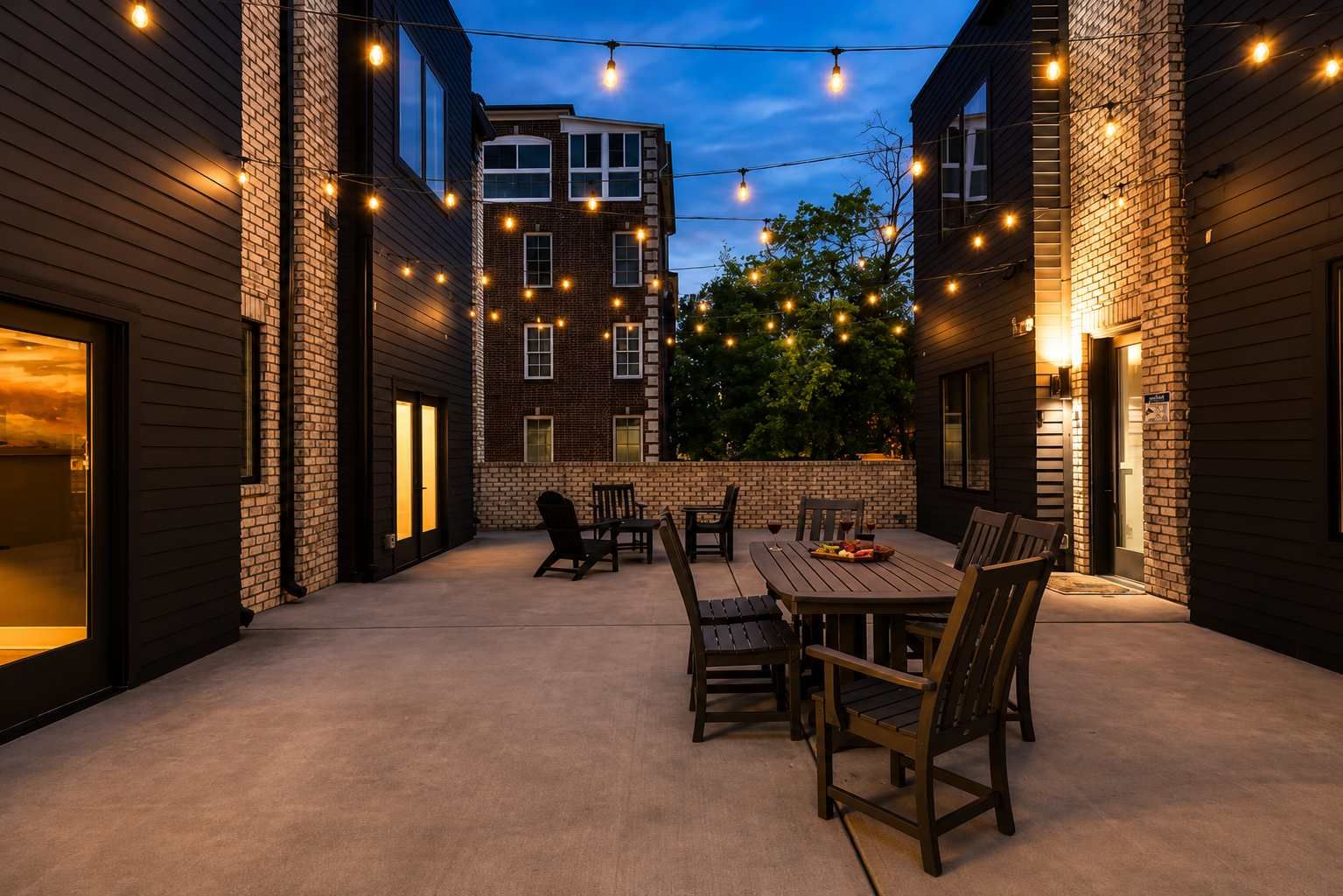 An outdoor patio with a wooden table and chairs, surrounded by brick walls and string lights.