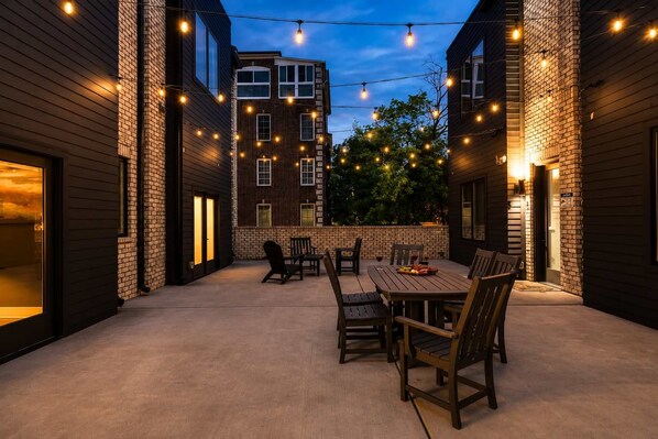 An outdoor patio with a wooden table and chairs, surrounded by brick walls and string lights.