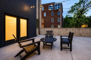 An outdoor patio with a round table and two chairs, a bench, and a building with a brick facade and a lit entrance.