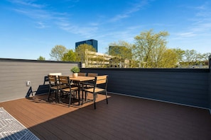 Una terraza al aire libre con una mesa y sillas de madera, una planta en maceta y vistas a edificios y árboles.