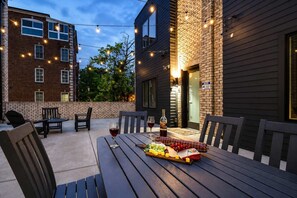 An outdoor patio with a table set for dining, surrounded by chairs, and a string of lights above. - New Build West End Oasis • Skyline Views (Nashville)