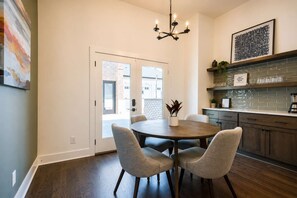 A modern kitchen with a round dining table, grey upholstered chairs, a chandelier, and a brick wall visible through the door. - New Build West End Oasis • Skyline Views (Nashville)