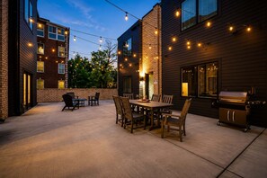 An outdoor patio with a dining table and chairs, a barbecue grill, and string lights.