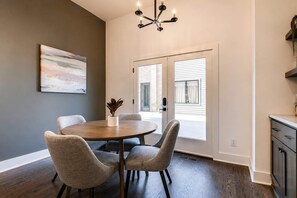 A dining area with a round table, four chairs, a chandelier, a painting, and a view through a door.