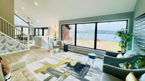 Amplia sala de estar con un gran ventanal que ofrece vistas a un lago, una escalera que sube a otro nivel y una decoración de estilo moderno.