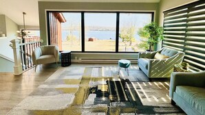 Amplia sala de estar con un ventanal que ofrece vistas a un lago y colinas. Hay dos sofás, una mesa de centro y una alfombra.