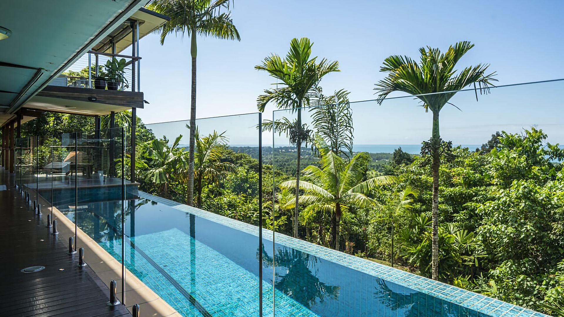 A clear view of a swimming pool with a glass barrier, surrounded by lush greenery and palm trees, overlooking a distant landscape.