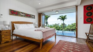A bedroom with a wooden bed, white bedding, a red and white patterned throw, a wooden nightstand with a radio, and a view of a balcony with palm trees. - Stu’s Lookout - Stunning Views (Wongaling Beach)
