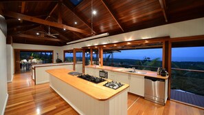 A modern kitchen with a central island, stainless steel appliances, and large windows offering a view of the landscape. - Stu’s Lookout - Stunning Views (Wongaling Beach)