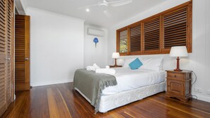 A bedroom with a bed, bedside tables, a ceiling fan, wooden shutters, and a wall-mounted air conditioner. - Stu’s Lookout - Stunning Views (Wongaling Beach)
