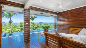 A balcony with a wooden floor, a bed with a white pillow, and a view of the ocean and greenery. - Stu’s Lookout - Stunning Views (Wongaling Beach)