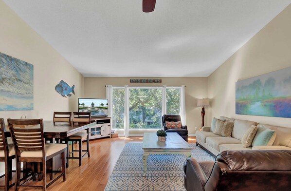 A spacious living room with a dining area, a large window with a view of trees, a flat-screen TV, and a sign reading "ISLE OF TALAS".