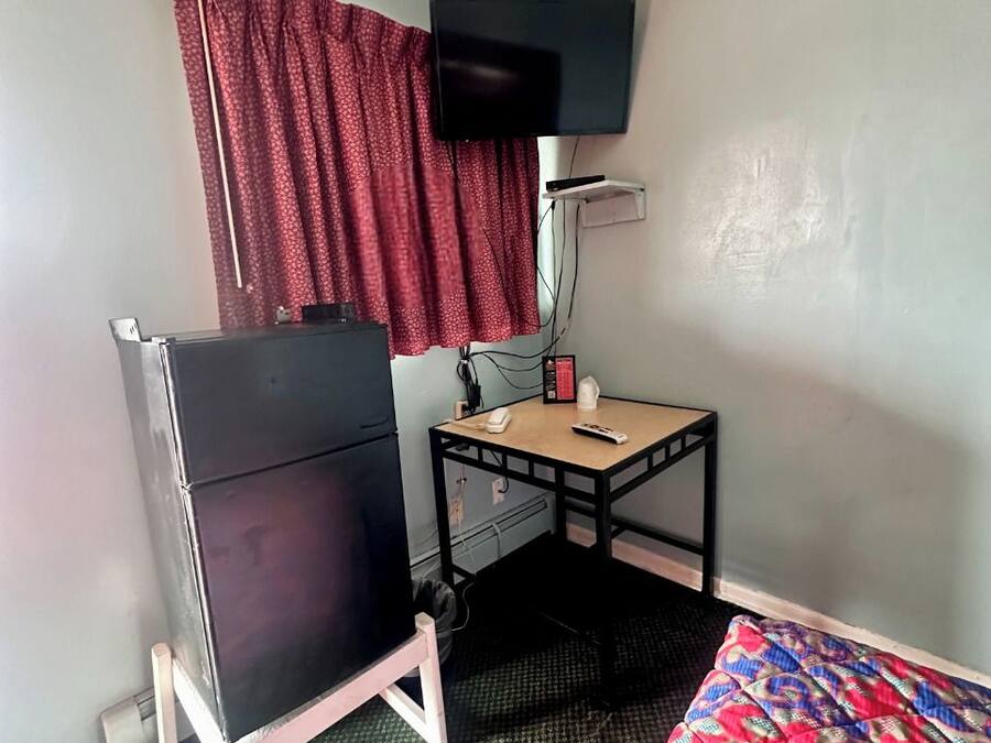 Quarto de hotel compacto com cama, pequena escrivaninha, televisão na parede e um refrigerador.