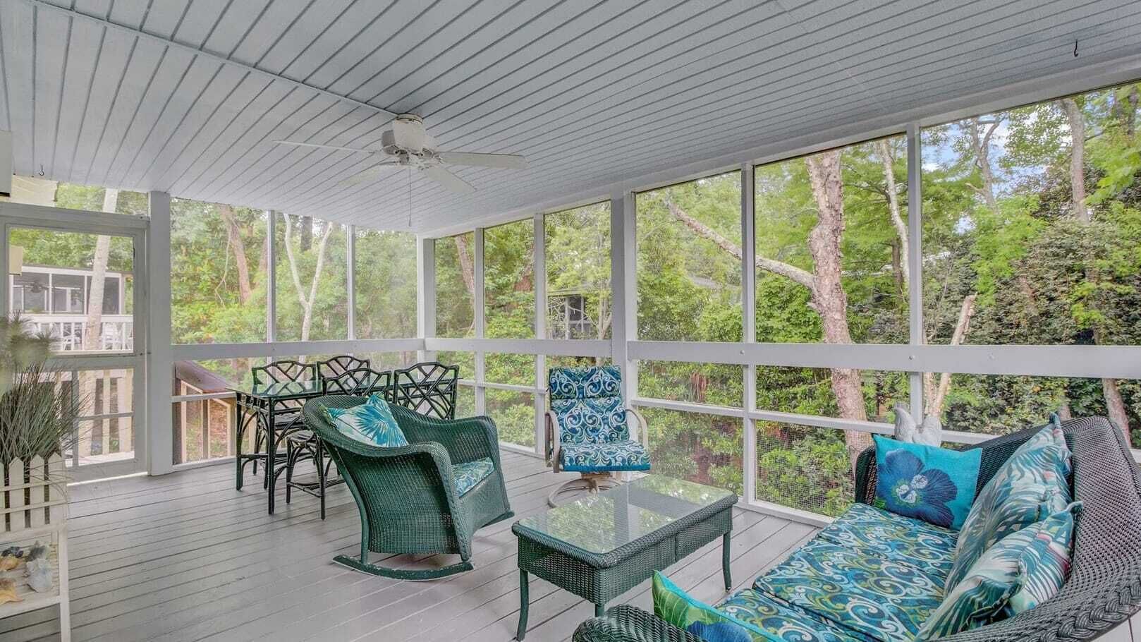 Un espace extérieur aménagé, abrité et doté de mobilier en osier et d’une table en verre, entouré de grandes fenêtres offrant une vue sur les arbres.