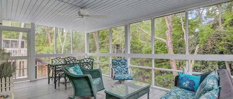 A covered outdoor seating area with wicker furniture and a glass table, surrounded by large windows offering a view of trees.