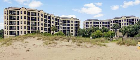 A beachfront hotel with multiple floors, balconies, and a sandy beach in the foreground.