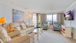 A spacious living room with a beige leather sofa, two armchairs, a glass coffee table, and a flat-screen TV mounted on the wall. - 311 Summerhouse by Avantstay Beach Views Access to Community Pool (Isle of Palms)
