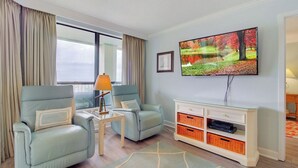 A living room with two light blue armchairs, a small table with a lamp, a glass cabinet with orange baskets, and a TV on the wall. - 311 Summerhouse by Avantstay Beach Views Access to Community Pool (Isle of Palms)