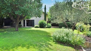 Um jardim bem cuidado, com diversas árvores e arbustos, um caminho bem definido e um edifício com porta e janelas amplas.