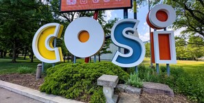 A sign with the word 'COSI' in large, colorful letters, surrounded by greenery and a stone pathway.