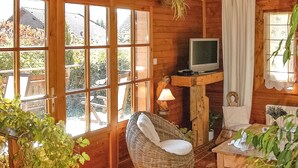 A cozy room with large windows, a wicker chair, a television on a wooden stand, and a small table with a lamp.