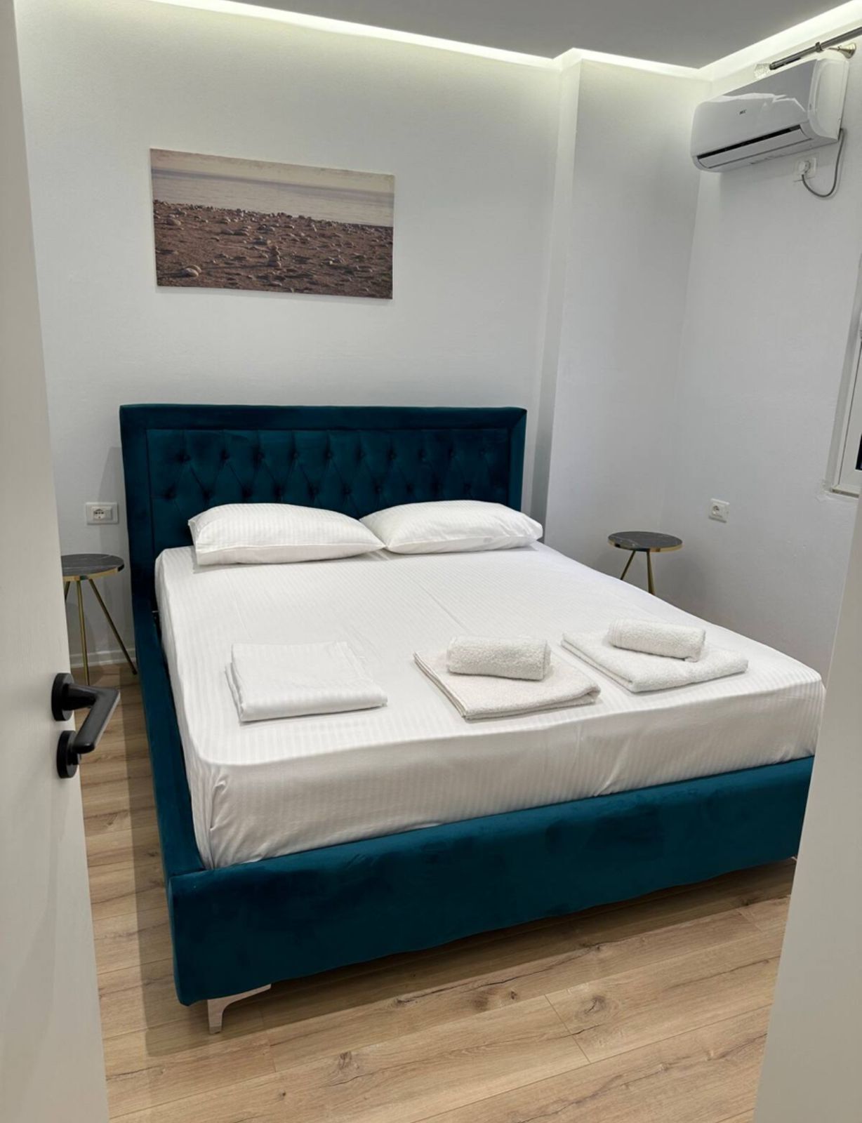 A neatly made bed with white linens and towels, a teal upholstered headboard, and a small bedside table with a lamp.