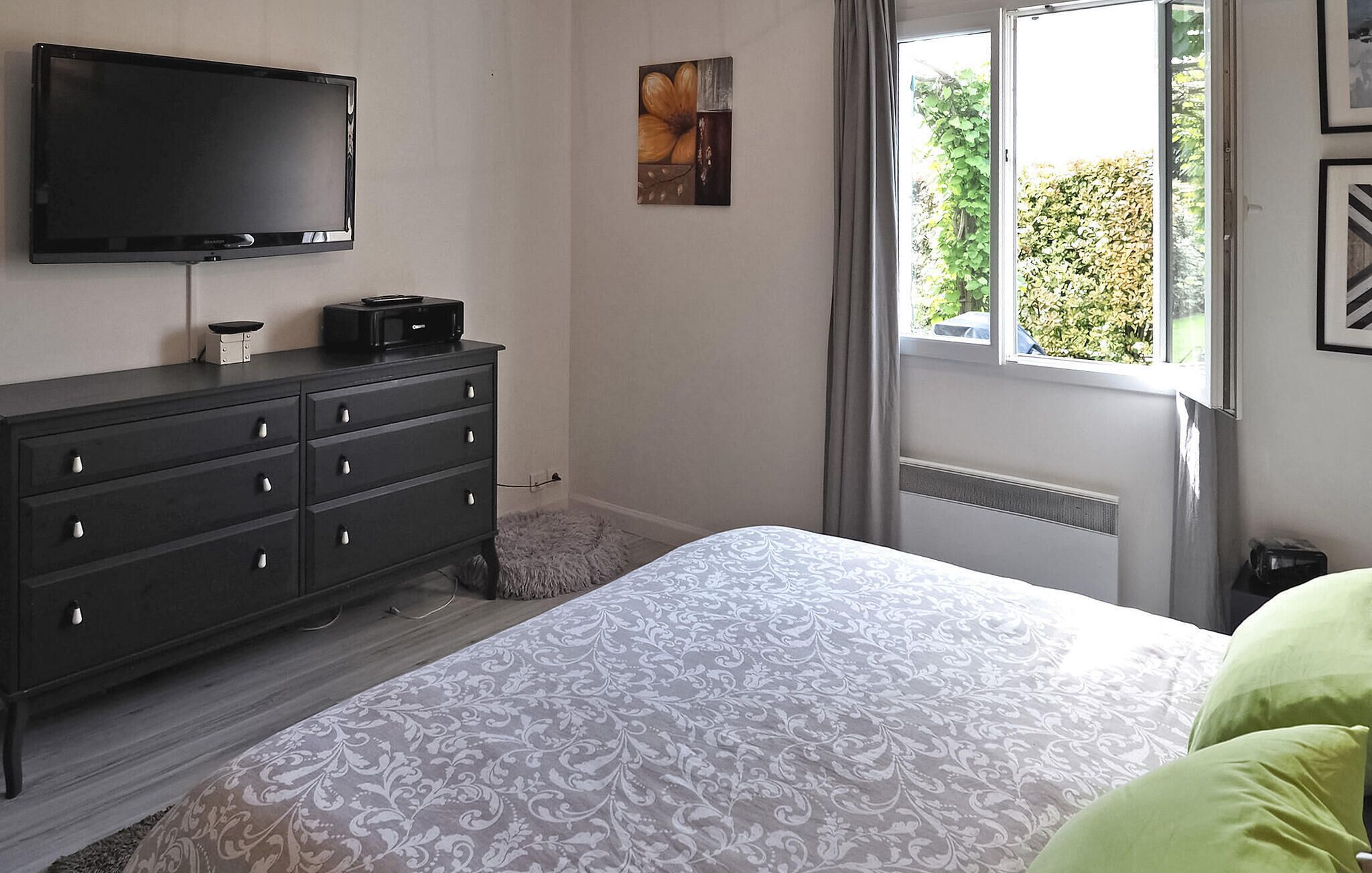 A bedroom with a bed, a dresser, a flat-screen TV, and a window with a view of greenery.