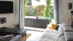 A modern living room with a grey sofa, a coffee table, and a flat-screen TV mounted on the wall. A sliding glass door leads to an outdoor area with a wicker table and a bowl of fruit.