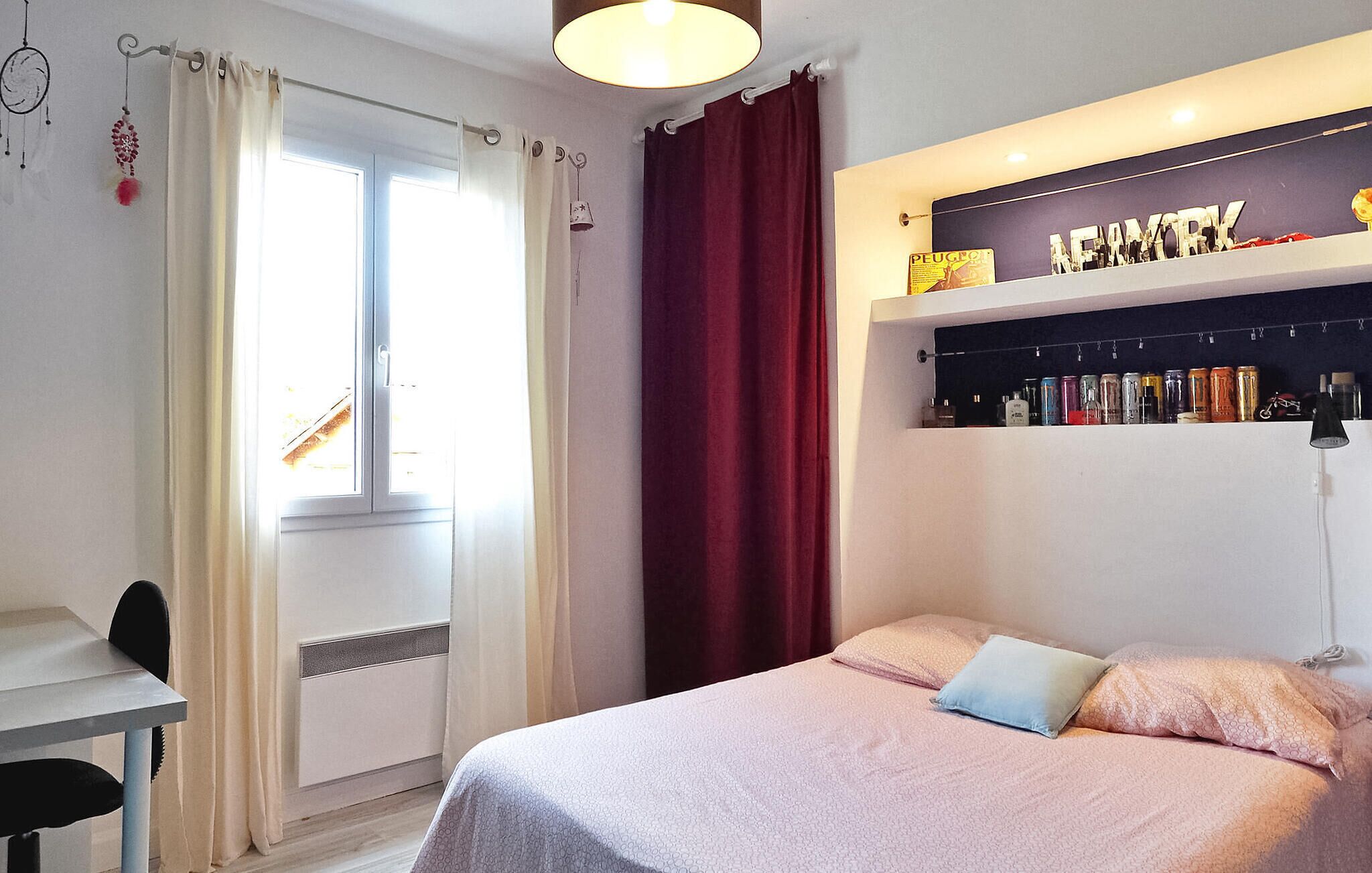 A bedroom with a bed, a desk, and a shelf with books and cans.