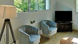 A living room with two light blue armchairs, a tripod floor lamp, a television on a wooden stand, and a large window with a view of trees. - Le Moulin De Lombraud (Brantome En Perigord)