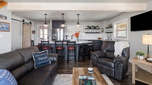 A modern living room with a sofa and armchair, a coffee table, and a kitchen area with bar stools. - Eagles Roost.  Wifi & Dog Friendly.  Covered porch & upper Deck w/ Sunset Views (Eagle Rock)