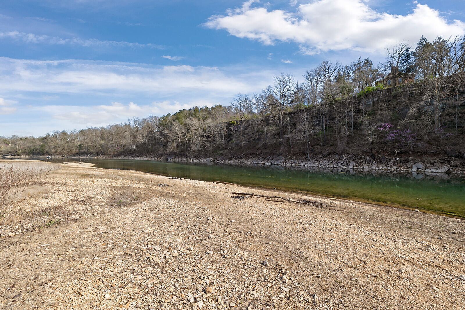 A riverbank with a rocky path leading to a house surrounded by trees.