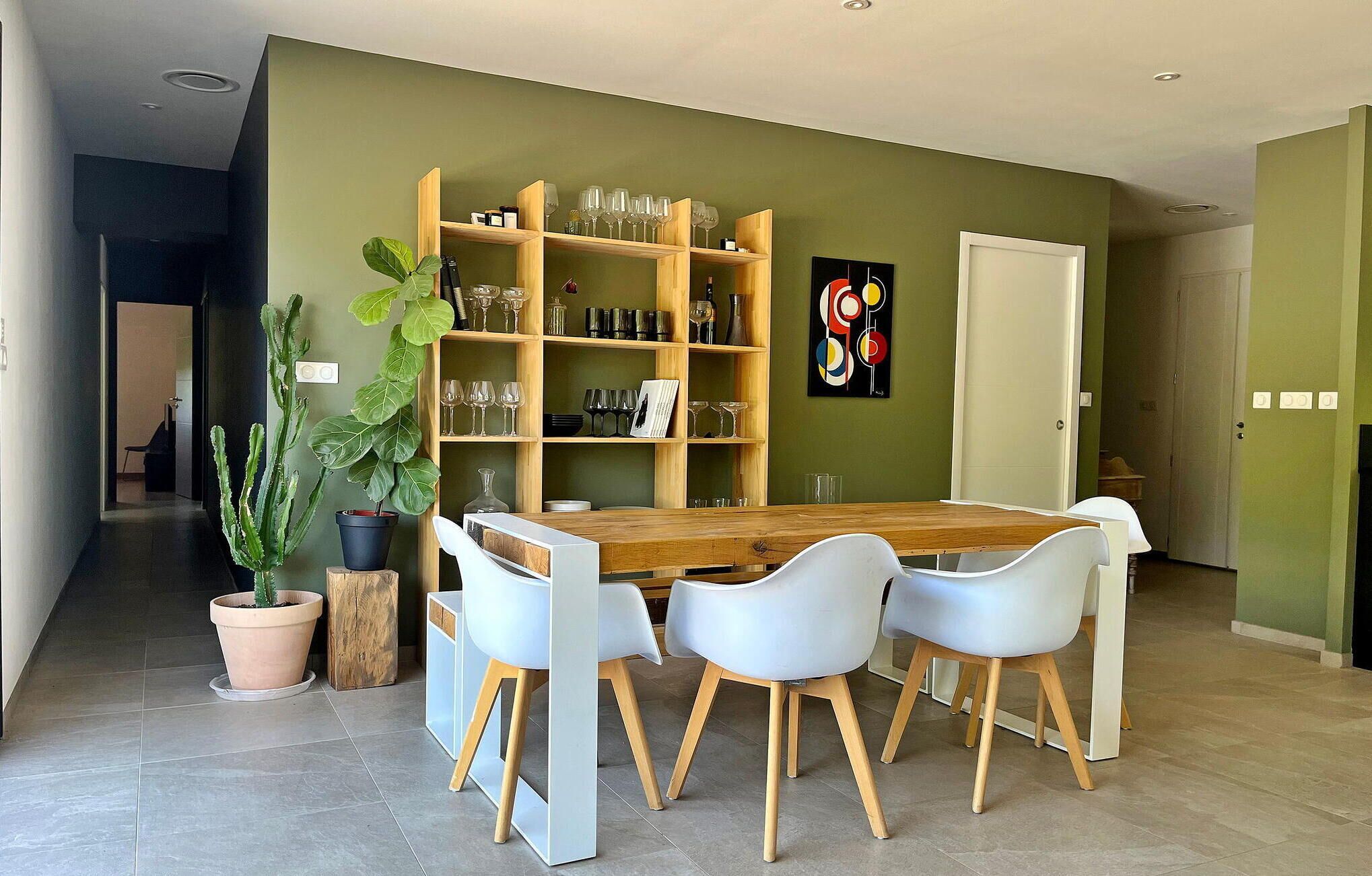 Un coin repas avec une table en bois et quatre chaises blanches, une bibliothèque avec des verres à vin et deux plantes en pot.
